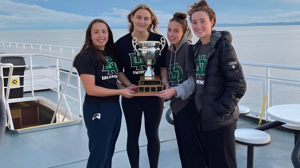 Un grupo de cuatro estudiantes de la Nanaimo District Secondary School sostiene un trofeo en la cubierta de un barco con agua y cielo nublado de fondo.