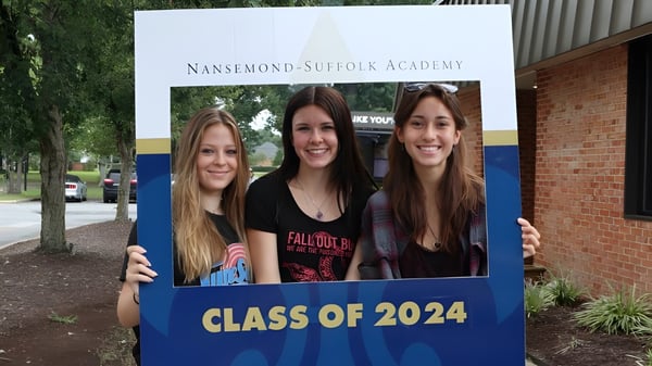 Tres estudiantes posan frente a un cartel de la Nansemond-Suffolk Academy con la inscripción Clase 2024.