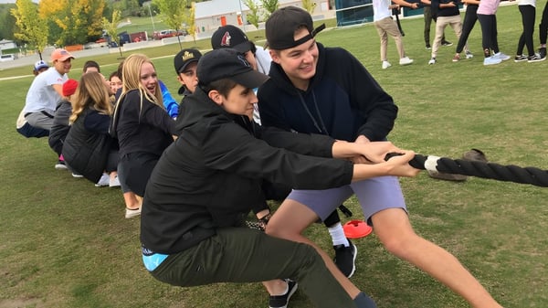 Un grupo de estudiantes de la National Sport School juega un partido en un campo deportivo con edificios y árboles de fondo.