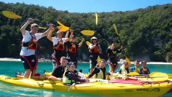 Estudiantes de Nayland College reman con chalecos salvavidas de colores y cascos en kayaks amarillos en aguas turquesas.
