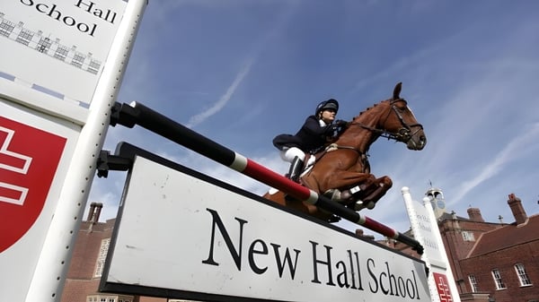Una estudiante de la New Hall School monta un caballo y salta sobre un obstáculo frente a un edificio escolar.