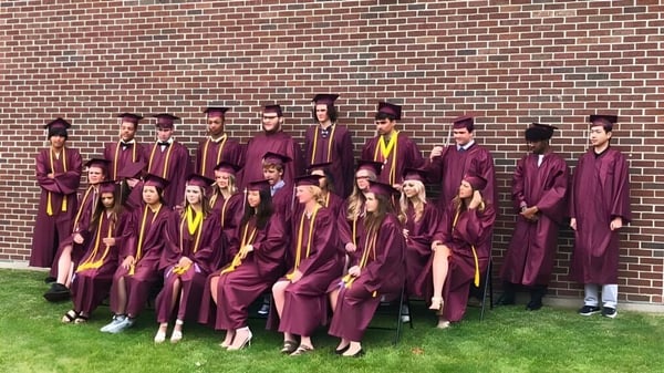 Graduadas y graduados de la N.E.W. Lutheran High School están en sus togas frente a una pared de ladrillo y celebran su graduación.