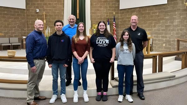 Estudiantes y adultos están de pie frente al edificio de ladrillo de la N.E.W. Lutheran High School con una bandera americana.