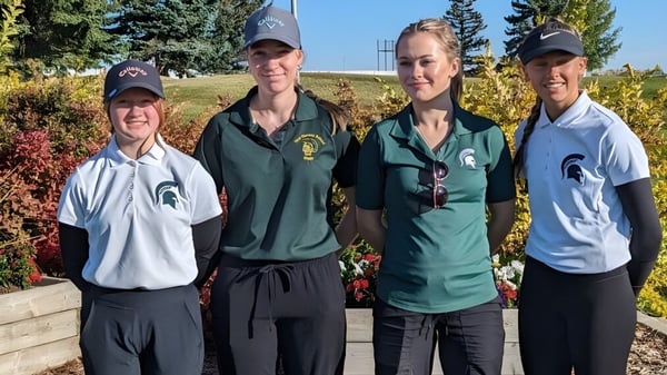 Cuatro estudiantes de la New Norway School en ropa deportiva de la Universidad Estatal de Michigan están frente a un fondo boscoso bajo un cielo azul claro.