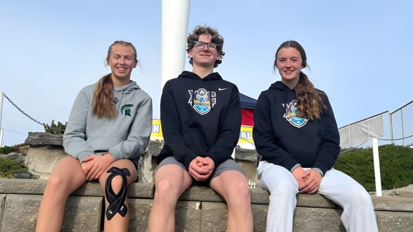 Tres estudiantes están sentados en un muro al aire libre frente a un cielo azul en el campus de la New Norway School.