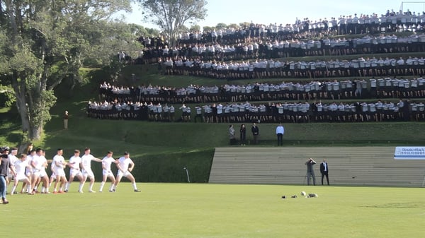 Un gran grupo de estudiantes de la New Plymouth Boys' High School se reúne en un prado frente a una alta estructura densa.