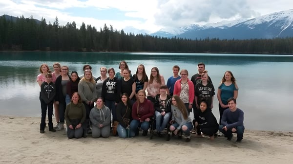 Estudiantes de la New Sarepta Community High School se reúnen en una playa de arena frente a un lago con montañas nevadas al fondo.