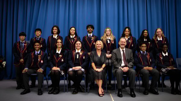 Un grupo de estudiantes en ropa formal está sentado frente a una cortina azul en el campus del Newlands College.