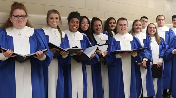 Un grupo de estudiantes de la Newman Central Catholic High School está en el pasillo con togas de graduación azules y camisas blancas.