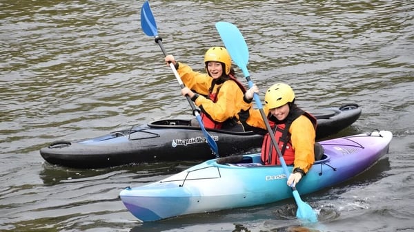 Un grupo de estudiantes del Newstead College rema con coloridos kayaks por un río a lo largo de una zona ribereña boscosa.