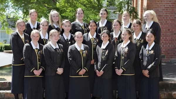 Un grupo de alumnas de la Nga Tawa Diocesan School está de pie en togas y birretes negros frente a un edificio de ladrillo con árboles.