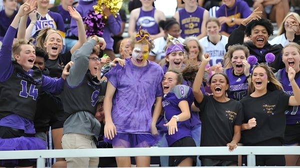 Un gran grupo de aficionados vestidos de morado y negro celebra en un evento deportivo de Norfolk Christian Schools.