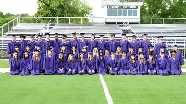 Un grupo de estudiantes de Norfolk Christian Schools está de pie en togas de graduación violetas en un campo frente a una estructura similar a un estadio.