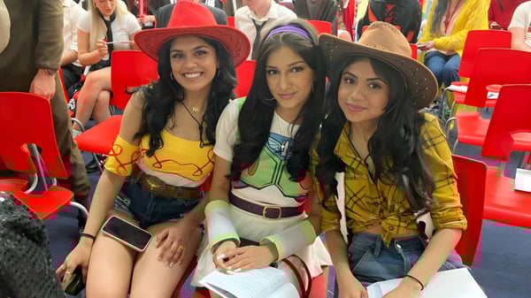 Tres estudiantes de la North Halifax Grammar School posan con atuendos coloridos frente a sillas rojas.