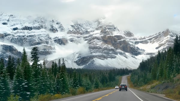 Una cadena montañosa cubierta de nieve se eleva sobre una carretera serpenteante a través de un valle boscoso cerca de la North Surrey Secondary School.