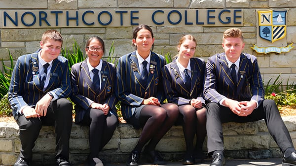 Un grupo de cinco estudiantes en uniforme escolar está sentado en un banco frente a un cartel del Northcote College.