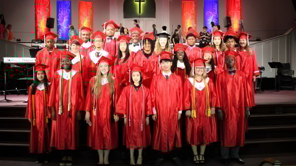 Un grupo de graduados de la Northside Christian Academy está de pie en togas rojas en una iglesia con una cruz de fondo.