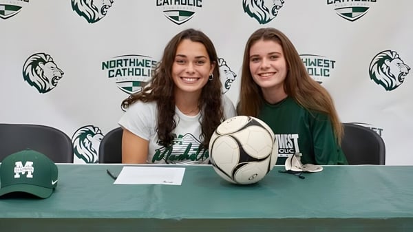 Dos estudiantes de Northwest Catholic High School están sentadas en una mesa con un balón de rugby frente a un fondo de logo.