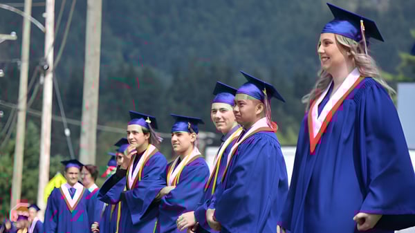 Un grupo de graduados de la Norwell District Secondary School está junto en togas de graduación azules frente a un fondo boscoso.