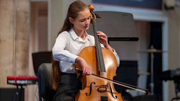 Una estudiante de la Norwich School toca el cello en una sala con paneles de madera e instrumentos musicales de fondo.