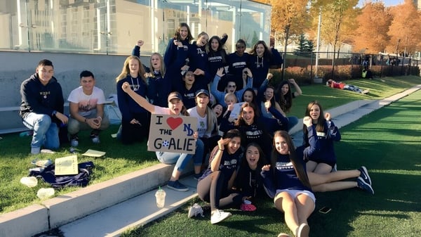Un grupo de estudiantes de la Notre Dame High School en Calgary se sienta y se pone de pie en un campo frente a un edificio escolar.