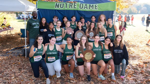 Un grupo de deportistas con camisetas verdes y blancas posan frente a un banner de Notre Dame High School en el bosque otoñal.