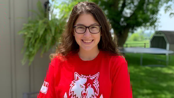 Una estudiante de la Nottawasaga Pines Secondary School sonríe frente a un edificio y un fondo de árboles verdes.