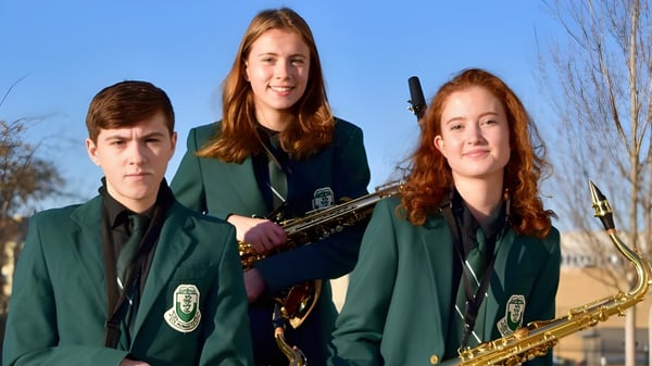 Tres estudiantes de la Oak Bay Secondary High School en uniformes verdes están con un saxofón y una trompeta bajo un cielo azul y árboles sin hojas.