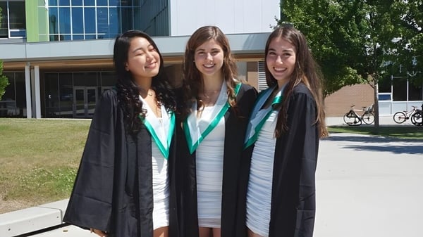Tres estudiantes de la Oak Bay Secondary High School están en togas de graduación frente a un edificio universitario con bicicletas y áreas verdes.