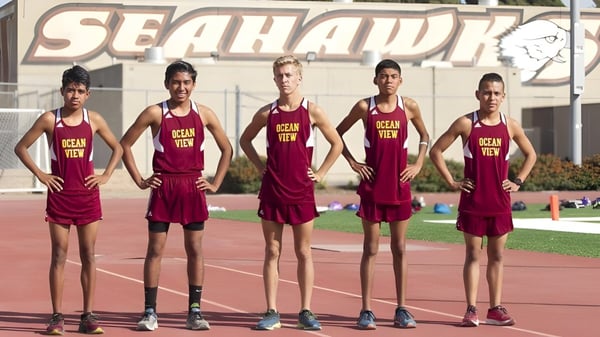 Cinco jóvenes atletas en camisetas rojas están en la pista deportiva de la Ocean View High School frente a un gran letrero de SEAHAWK.