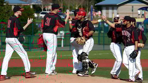 Un grupo de jugadores de béisbol del Olympic College celebra en el campo con un edificio y árboles de fondo.