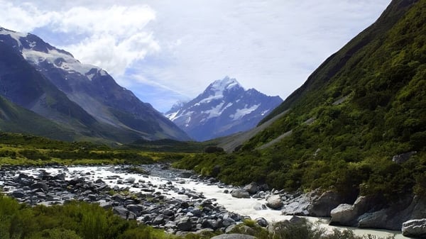 Una cadena montañosa nevada se ve detrás de vegetación verde y un arroyo en el terreno de Onslow College.
