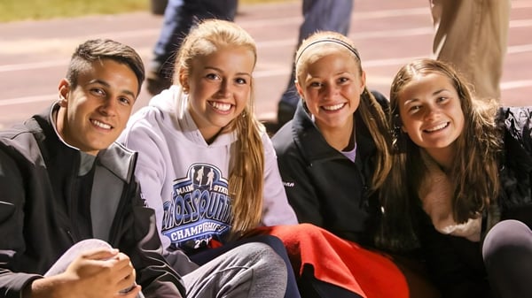 Un grupo de cuatro estudiantes de la Orono High School se sienta riendo en un campo deportivo frente a un edificio de ladrillo.