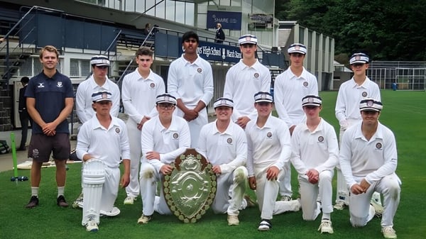 Un grupo de estudiantes de la Otago Boys' High School posan en un campo de cricket con un estadio al fondo.