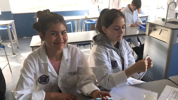 Dos estudiantes en el laboratorio del Our Lady Catholic College llevan batas de laboratorio blancas y trabajan con equipo médico.
