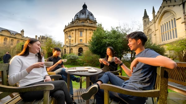 Un grupo de estudiantes conversa y disfruta de bebidas en el área exterior del Oxford International College frente a un edificio histórico.