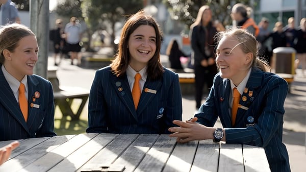 Tres estudiantes en uniformes escolares están sentadas en una mesa de madera y conversan en el campus del Papamoa College.