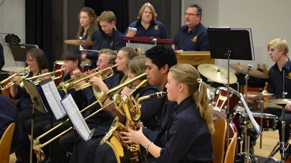 Un grupo de músicos de Papanui High School en uniformes azul oscuro toca en un escenario varios instrumentos de viento y de madera.