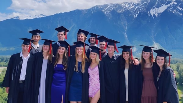 Un grupo de graduados está de pie con ropa académica frente a un paisaje montañoso en el terreno de la Pemberton Secondary School.