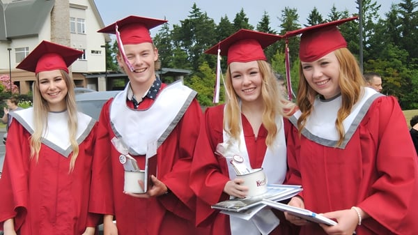 Un grupo de graduadas y graduados de Pender Harbour Secondary está afuera con togas rojas y birretes frente a un fondo boscoso.