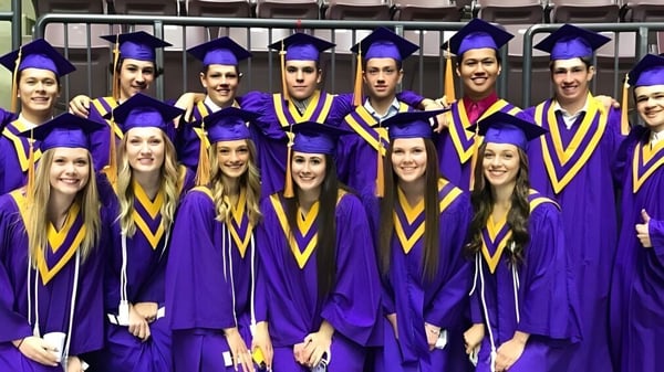 Graduadas y graduados de la Penticton Secondary School están en togas violetas y doradas para una foto en un interior.