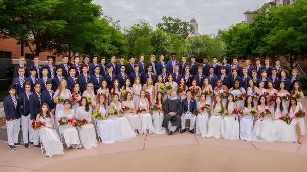 Un gran grupo de personas vestidas de manera formal se reúne al aire libre frente a un edificio con una torre alta en el campus de la Perkiomen School.