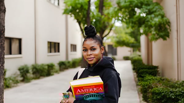 Una joven con un peinado llamativo camina por una calle bordeada de árboles llevando un libro en el campus de la Phoenix Christian Preparatory School.