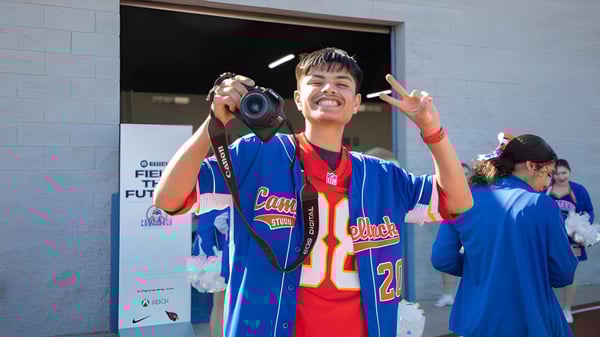 Un joven en camiseta deportiva azul sostiene una cámara y muestra un signo de paz frente a un edificio del Phoenix Union High School District.