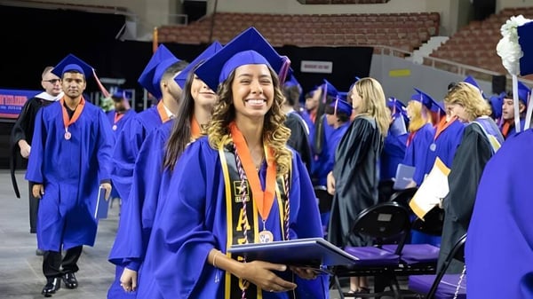 Un grupo de graduados del Phoenix Union High School District está en una ceremonia de graduación en interiores con togas azules y diplomas.
