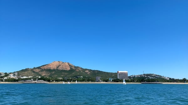 Una cima rocosa de montaña se eleva sobre un lago tranquilo con edificios al fondo en el terreno de la Pimlico State High School.
