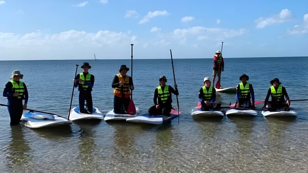 Estudiantes del Plympton International College están con chalecos salvavidas en tablas de paddle en el agua poco profunda frente a un océano tranquilo.