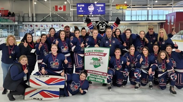 El equipo femenino de hockey sobre hielo de la Polyvalente Thomas-Albert está en sus camisetas azules en el pabellón de hielo frente a la bandera canadiense y británica.