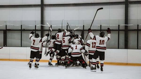 Un grupo de jugadores de hockey de la Pomfret School se reúne sobre el hielo de una pista de patinaje frente a una pared de vidrio.