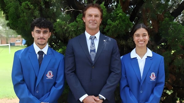 Tres personas, incluyendo a dos alumnos en uniformes azules, están de pie frente a un fondo verde en el terreno de la Port Hacking High School.